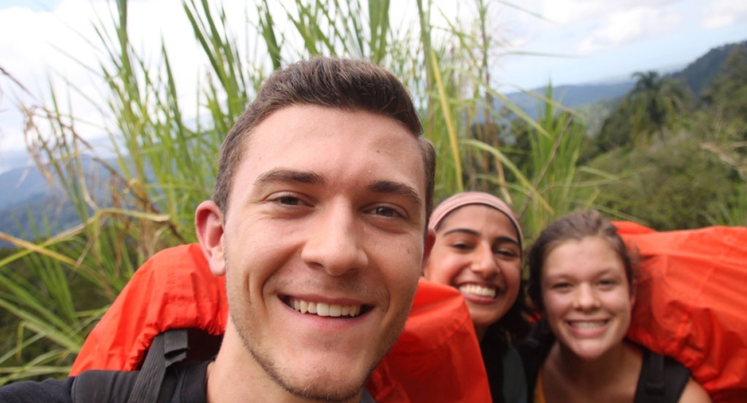 Three people smile while standing in front of a green landscape. Two of them are wearing orange backpacks. 